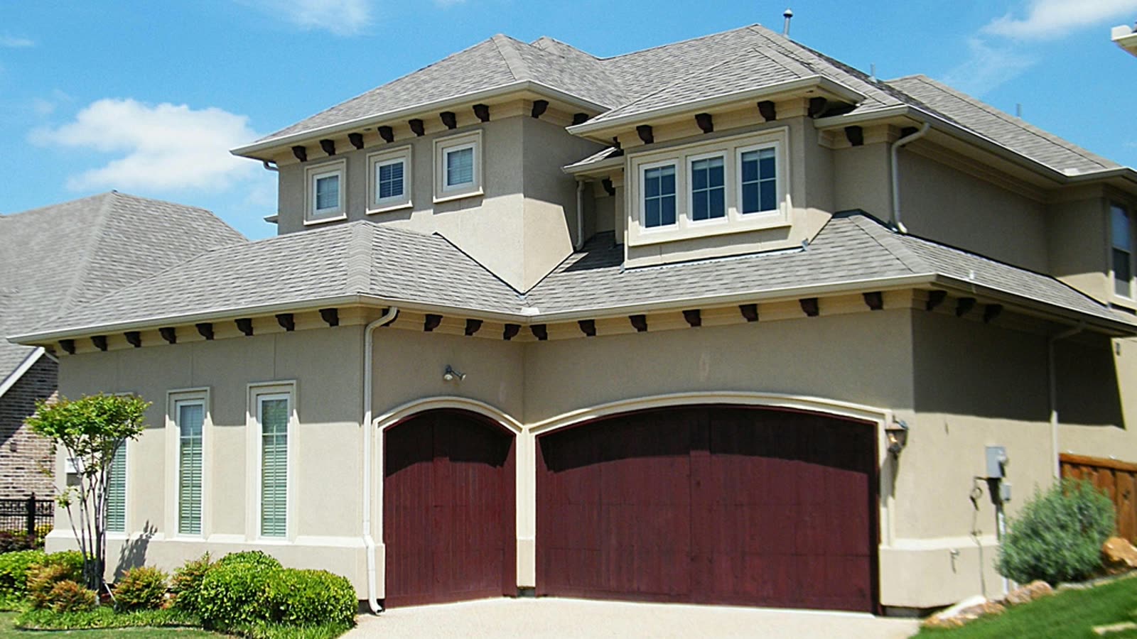 A two-story Texas suburban house with beige stucco, a hipped composition-shingle roof with exposed rafter tails, and two arched dark-red wood garage doors facing a concrete driveway